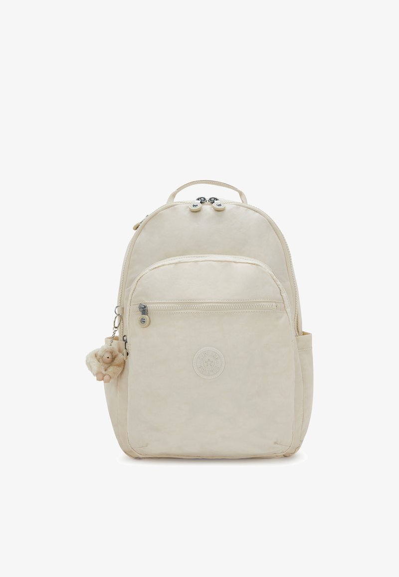 Beige reppu, jossa on pyöreä muoto, kangasmateriaali, kaksoispainike suljenta, etutasku, sivutasku ja pörröinen apina avaimenperi, joka on kiinnitetty.