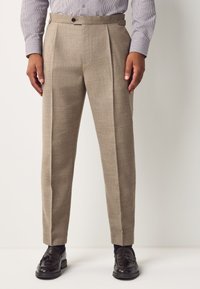 Beige színű, texturált felületű, fazonra szabott nadrág, elöl redőkkel, övbújtatókkal és egy gombos záródással, fekete mokaszin cipővel párosítva.