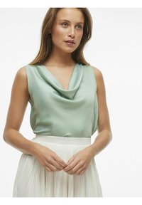 Mätová hodvábna bezrukávová blúzka s drapovaným výstrihom, v kombinácii s plisovanou bielou sukňou. Hladká textúra, minimalistický dizajn.