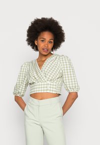 Blusa cropped de gingham verde e branco com mangas bufantes, apresentando um design envolvente, laterais franzidas e detalhes de botões. Combinada com calças verde claro.