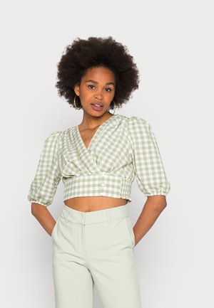 Blusa cropped de gingham verde e branco com mangas bufantes, apresentando um design envolvente, laterais franzidas e detalhes de botões. Combinada com calças verde claro.