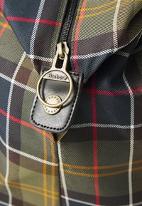 Close-up of a Barbour zipper pull on tartan-patterned fabric with green, navy, red, white, and yellow lines.
