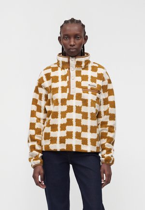 Personne portant un pull polaire à carreaux beige et blanc avec des boutons et une poche poitrine, debout devant un fond uni.