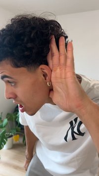 Joven con cabello rizado que lleva una camiseta blanca. Pendiente de aro dorado en la oreja izquierda. Mano cerca de la oreja, expresión concentrada, ambiente interior.