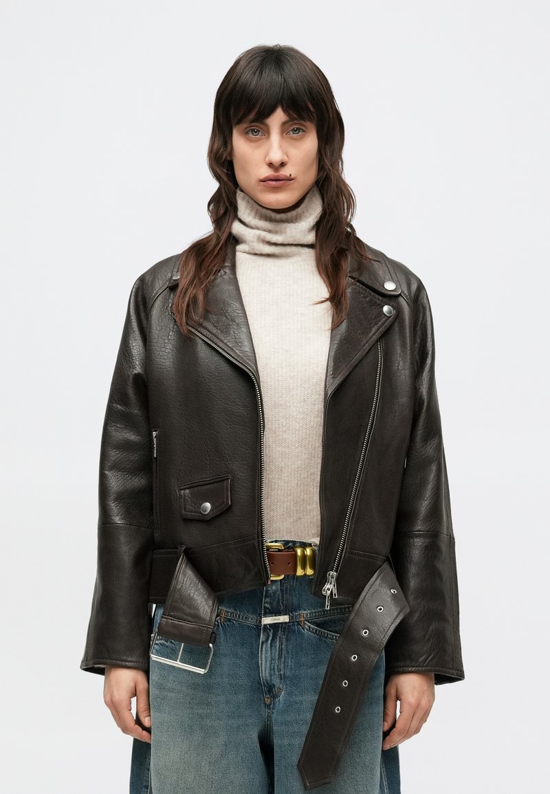 Zwarte leren motorjack met ritssluiting aan de voorkant, zijzakken en decoratieve details. Gedragen over een lichte coltrui en blauwe spijkerbroek.