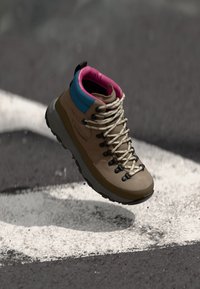 Botte de randonnée brune avec un col bleu et rose, dotée de lacets beiges, d'œillets noirs et d'une semelle grise texturée. Flottant au-dessus d'une ligne blanche.