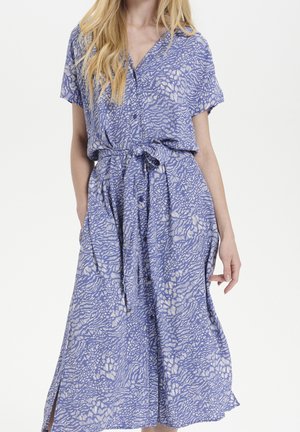 Donna che indossa un vestito a maniche corte con motivo blu e bianco, con bottoni e cintura annodata in vita, in piedi davanti a uno sfondo semplice.