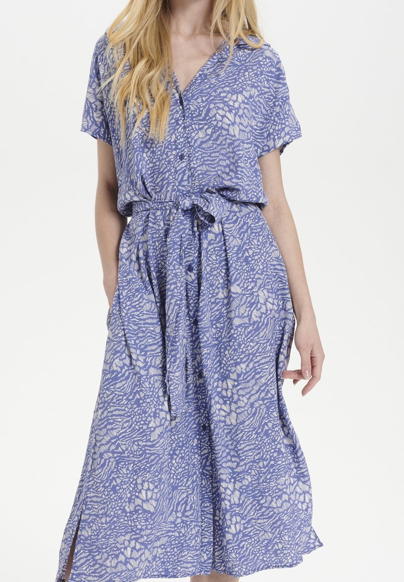 Femme portant une robe à manches courtes bleue et blanche à motifs, avec des boutons et une ceinture nouée à la taille, debout devant un fond uni.