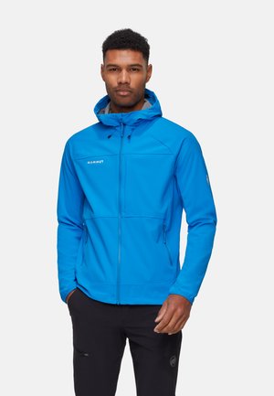 Uomo che indossa una giacca blu Mammut con cappuccio e cerniera, con le mani nelle tasche, in piedi davanti a uno sfondo chiaro e uniforme.