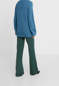 Pull en maille côtelée bleu à manches longues, associé à un pantalon vert à jambes larges avec un motif géométrique. Chaussures noires visibles.