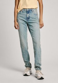 Gai zilu džinsi ar augsto jostasvietu, taisnu kāju un viegli izbalējušu izskatu. Saskaņoti ar neitrālu toņu kedām un dzeltenu blūzi.