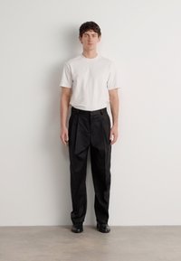 Weißes T-Shirt mit kurzen Ärmeln, kombiniert mit hochgeschnittenen schwarzen Faltenhosen und schwarzen Schuhen, stehend vor einem schlichten weißen Hintergrund.