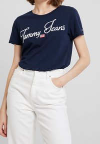 Tmavě modré bavlněné tričko s bílou vyšívanou logem "Tommy Jeans" a malým detailem americké vlajky. Kombinováno s bílými džíny s vysokým pasem.