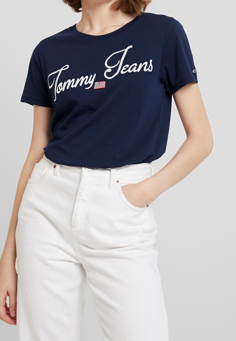 Tmavě modré bavlněné tričko s bílou vyšívanou logem "Tommy Jeans" a malým detailem americké vlajky. Kombinováno s bílými džíny s vysokým pasem.
