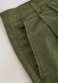 Nærbillede af olivengrønne bukser, der viser linningen, bæltestrop og sidelomme med detaljerede sømme.