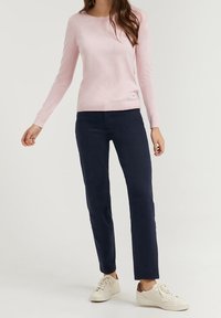 Light pink sweater with long sleeves, fitted design, and ribbed cuffs, paired with navy pants and white sneakers. Notable brand tag displayed.