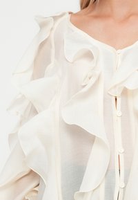 Gjennomsiktig hvit bluse med rynkede detaljer langs utringningen og knapper foran, laget av lett stoff.