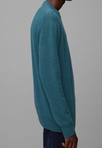 Tyrkysový pletený svetr s kulatým výstřihem, dlouhými rukávy a texturovaným povrchem, doplněný tmavými kalhotami.