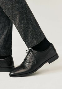 Mustat nahkakengät brogue-tyylillä, joissa on rei'itettyä yksityiskohtaa, terävä kärki ja matala korko, yhdistettynä tummiin räätälöityihin housuihin ja sukkiin.