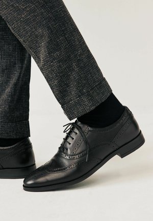 Zapato brogue de cuero negro con detalles perforados, punta afilada y tacón bajo, combinado con pantalones oscuros de corte ajustado y calcetines.