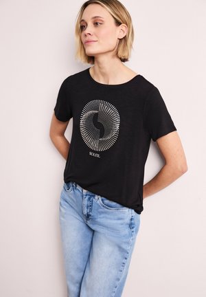 Kobieta o krótkich blond włosach, ubrana w czarną koszulkę z białym wzorem spiralnych kropek i napisem "SOLEIL", w połączeniu z jasnoniebieskimi dżinsami.