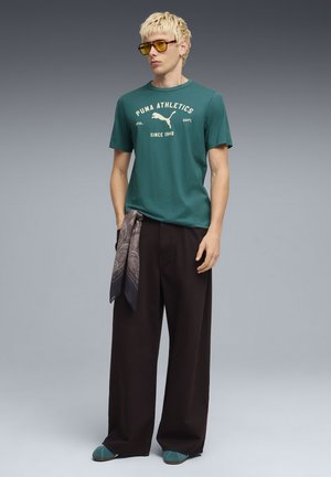 Maglietta di cotone verde con stampa "PUMA ATHLETICS", pantaloni neri larghi, sciarpa grigia con motivo e scarpe slip-on turchesi con suola piatta.