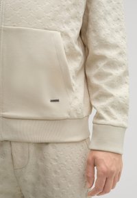 Giacca zip-up beige chiaro con motivo floreale testurizzato, tasca frontale e orlo a costine. Pantaloni abbinati con dettagli di design simili.