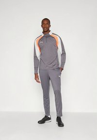 Szürke melegítő narancssárga és fehér részletekkel, hosszú ujjakkal, cipzáras gallérral és testhezálló nadrággal. Az anyag simának tűnik; a modell fekete cipőt visel.