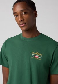 Groene katoenen t-shirt met korte mouwen. Kenmerkt zich door een meerkleurige geborduurde logo-tekst op de borst. Ronde halslijn en klassieke pasvorm.