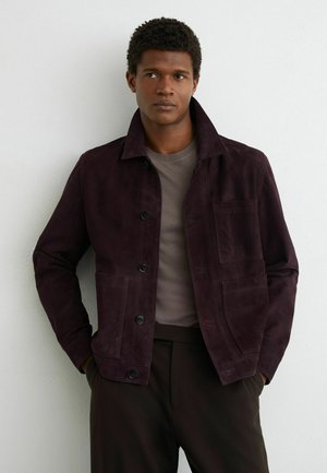 Man with short curly hair wearing a dark purple buttoned jacket over a gray shirt, standing with hands in pockets against a white wall.