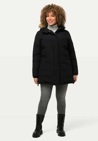 Svart puffjacka med en huva fodrad med fuskpäls, framdragkedja och sidofickor, i kombination med grå jeans och svarta snörstövlar.