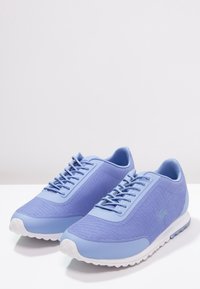Světle modré síťované běžecké boty s plochými tkaničkami, texturovaným povrchem, zaoblenou špičkou a bílou gumovou podrážkou. Logo Lacoste na boční straně.