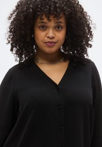 Μαύρο πουκάμισο με κουμπιά, V λαιμόκοψη, υφασμένο ύφασμα και κοντά μανίκια. Διαθέτει διακριτικές κάθετες ρίγες και στρογγυλά μαύρα κουμπιά.