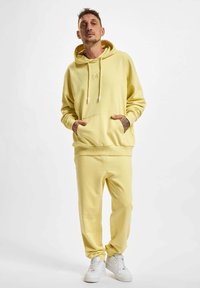 Gelbes Kapuzenoberteil mit Kordeln und Fronttasche, kombiniert mit passenden gelben Sweatpants, getragen mit weißen Sneakers. Weicher, lässiger Stoff.
