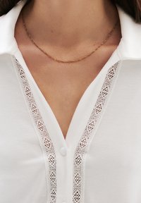 Witte blouse met een tekstuur van kant langs de knoopsluiting, met een knoopsluiting. Een dunne, gedraaide gouden ketting is gedragen.