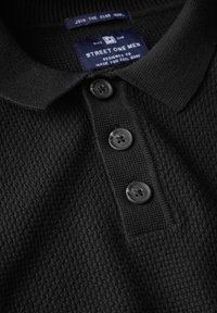 Svart strukturerad polo med krage, treknapps placket med mörka knappar och en vävd etikett med varumärkesinformation.