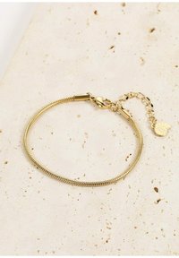 Bracciale in metallo dorato con design liscio e flessibile. Presenta una chiusura a moschettone e una catena di estensione con un piccolo charm. Superficie testurizzata.