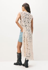 Helebeežis heegeldatud pikk kampsun lahtise lõike ja avatud külgedega, mis on kombineeritud teksaseelikuga ja mustade põlveni ulatuvate kontsakingadega.