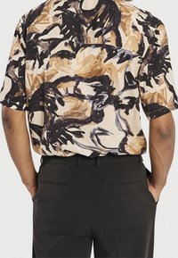 Lyhythihainen paita, jonka pohjaväri on kerma ja jossa on abstrakteja ruskeita ja mustia kuvioita. Yhdistetty tummien housujen kanssa. Asun takaosa.