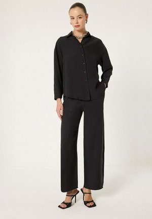 Γυναίκα που φοράει μαύρο πουκάμισο με κουμπιά και μακριά μανίκια, φαρδιά μαύρη παντελόνια, μαύρα ψηλοτάκουνα σανδάλια και χοντρό ασημένιο κολιέ.