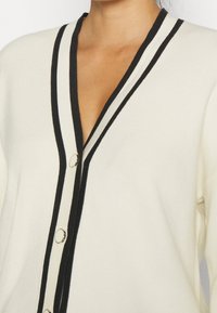 Krémový cardigan s černými pruhy, s designem do V, ribbovanou texturou a stříbrnými knoflíkovými akcenty po celé přední části.