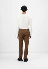 Man met krullend haar staat met zijn gezicht naar een witte muur, gekleed in een wit shirt met lange mouwen, bruine broek en zwarte schoenen.