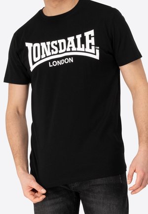 Mann, der ein schwarzes Lonsdale London T-Shirt und dunkle Jeans trägt, steht mit entspannten Armen vor weißem Hintergrund.
