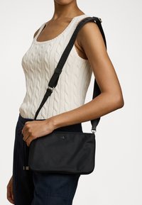 Sac à bandoulière noir avec une texture lisse, de forme rectangulaire slim, une sangle ajustable et un discret logo sur le devant.