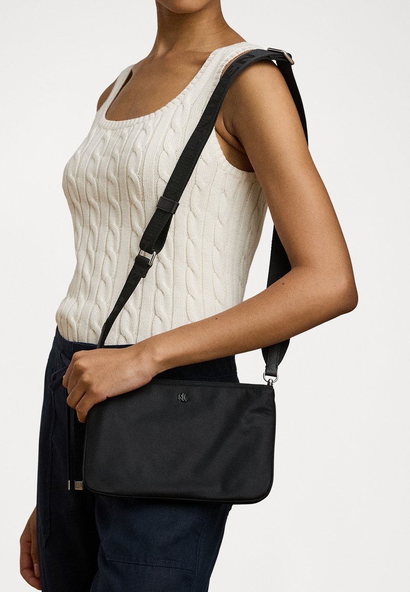 Sac à bandoulière noir avec une texture lisse, de forme rectangulaire slim, une sangle ajustable et un discret logo sur le devant.