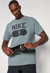 Nike T-shirt i falmet blå med stort sort logo og nummer 1, samlet med en sort isoleret vandflaske, hænder der holder flasken.