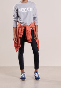 Grauer Sweatshirt mit "HUGO" in Weiß gedruckt, orangefarbene Jacke um die Taille gebunden, schwarze Leggings und blaue Sneaker mit weißen Schnürsenkeln.