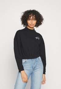 Sudadera negra recortada con cuello alto, puños y cinturilla acanalados; cuenta con bordado del logotipo blanco en el pecho. Llevada con vaqueros de color azul claro.