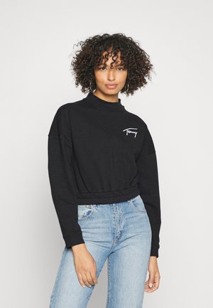 Felpa nera cropped con collo alto, polsini e vita a coste; presenta un ricamo bianco del logo sul petto. Indossata con jeans blu chiaro.