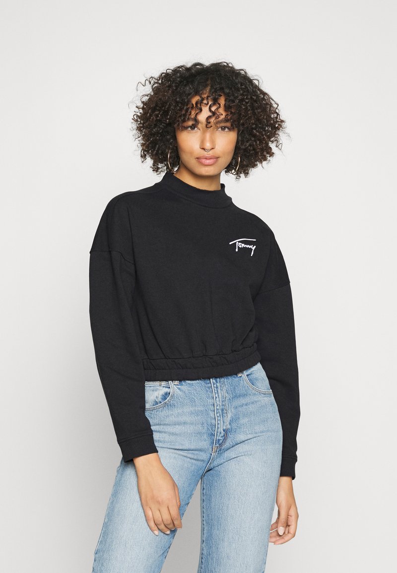 Sudadera negra recortada con cuello alto, puños y cinturilla acanalados; cuenta con bordado del logotipo blanco en el pecho. Llevada con vaqueros de color azul claro.